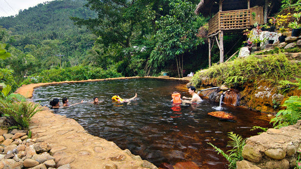People enjoying a natural pool surrounded by lush greenery and a thatched-roof structure.